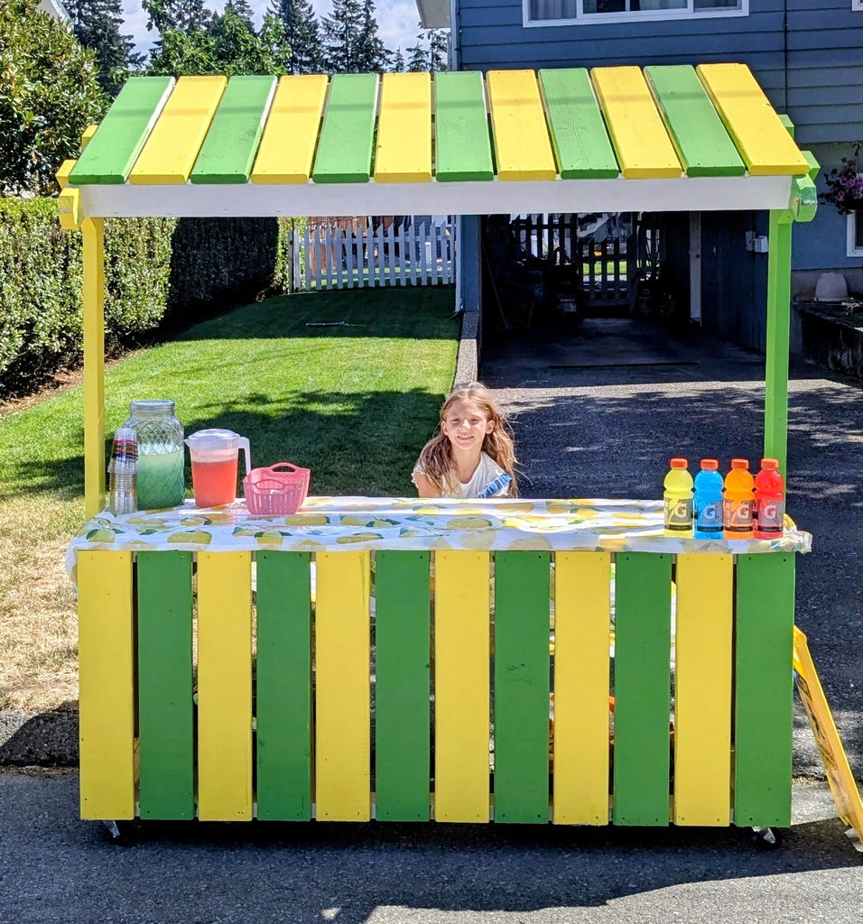 Girl sitting at a wooden, yellow and green lemonade stand at end of driveway.