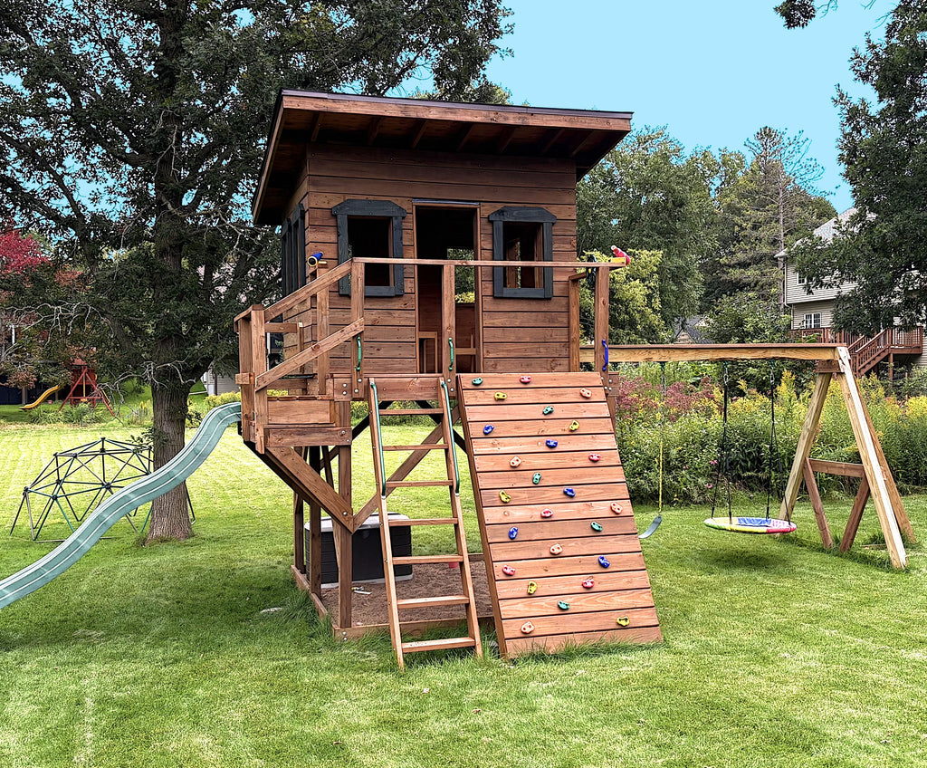 Lean-to roof kid's wooden playhouse on 4x4x posts with rock wall, swingset, and wavy slide with step-down platform in a back yard.