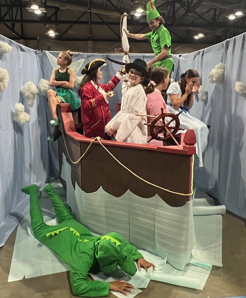 Children in costumes on a pretend pirate ship with a person in a green dinosaur costume on the ground.