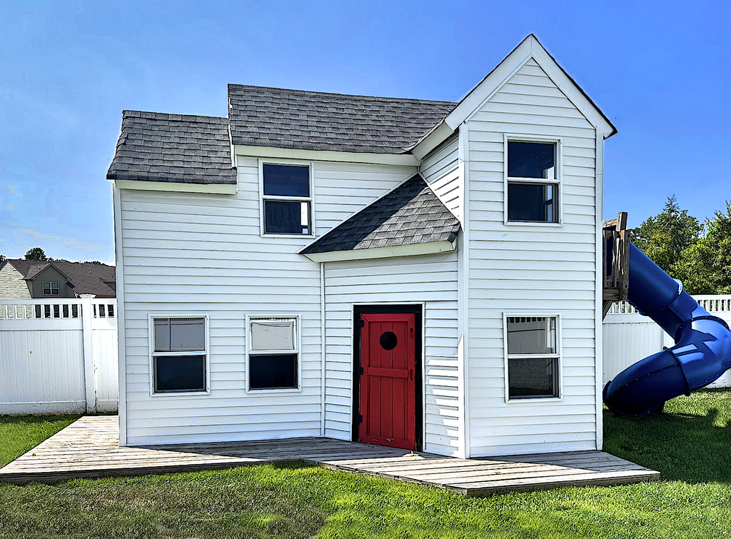 Large white, two story playhouse with gable roof, red front door and blue spiral slide in a backyard setting.