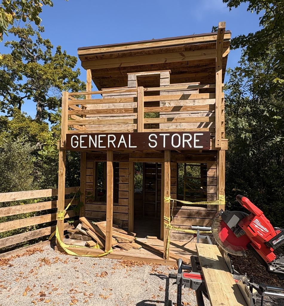 Wooden playhouse labeled 'General Store' with tools and materials around, set against a natural background.