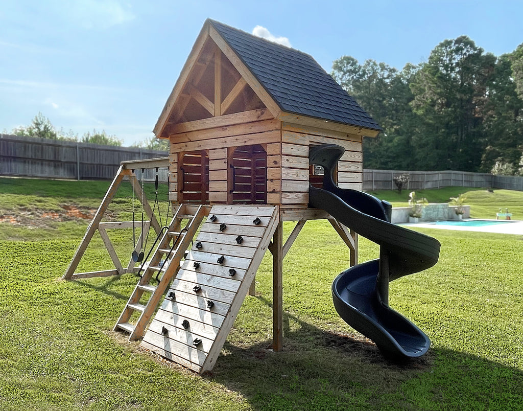 Open gable, wooden playhouse with a spiral green slide in a backyard with a fence and pool.
