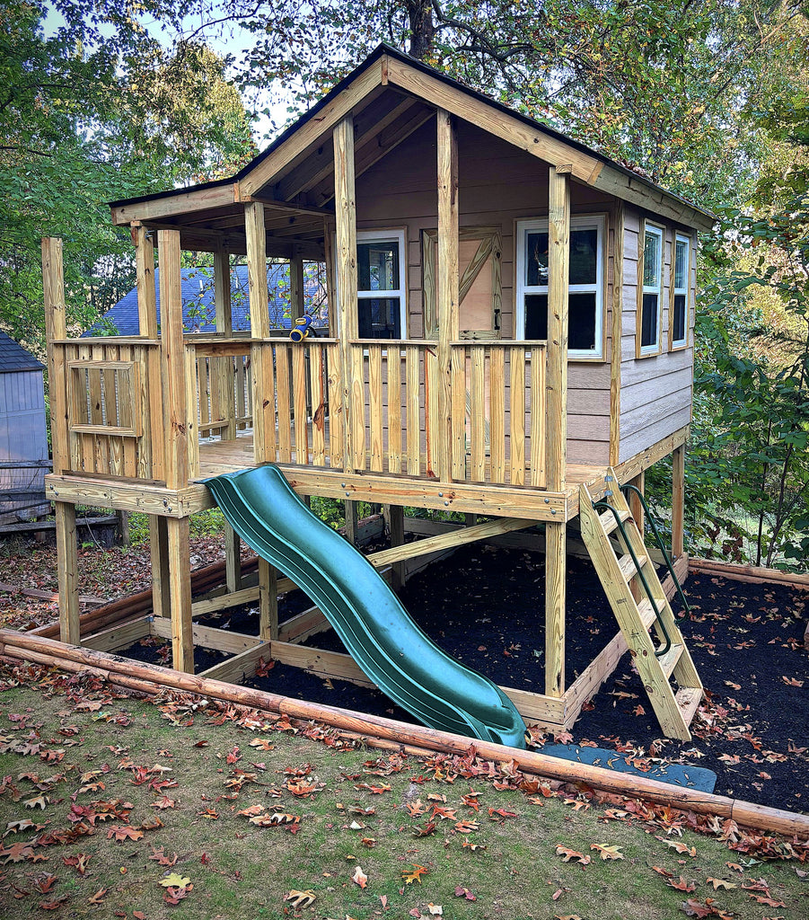 Wooden playhouse with a green slide on a grassy area with trees in the background