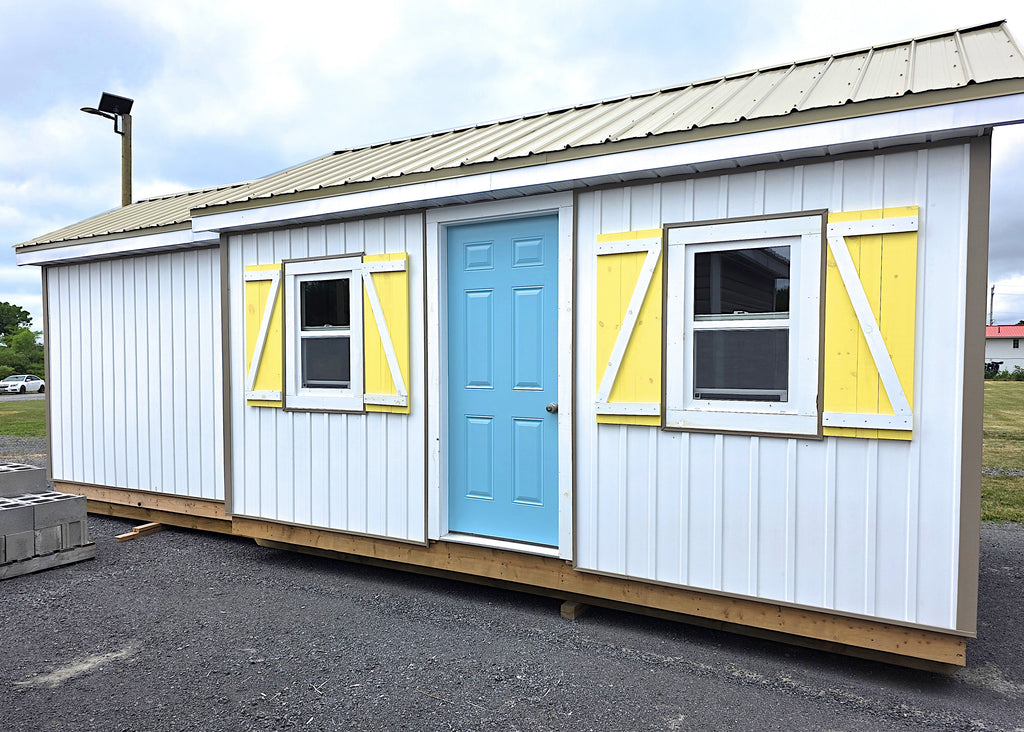 A white shed with blue door with metal gable roof and bump-out.