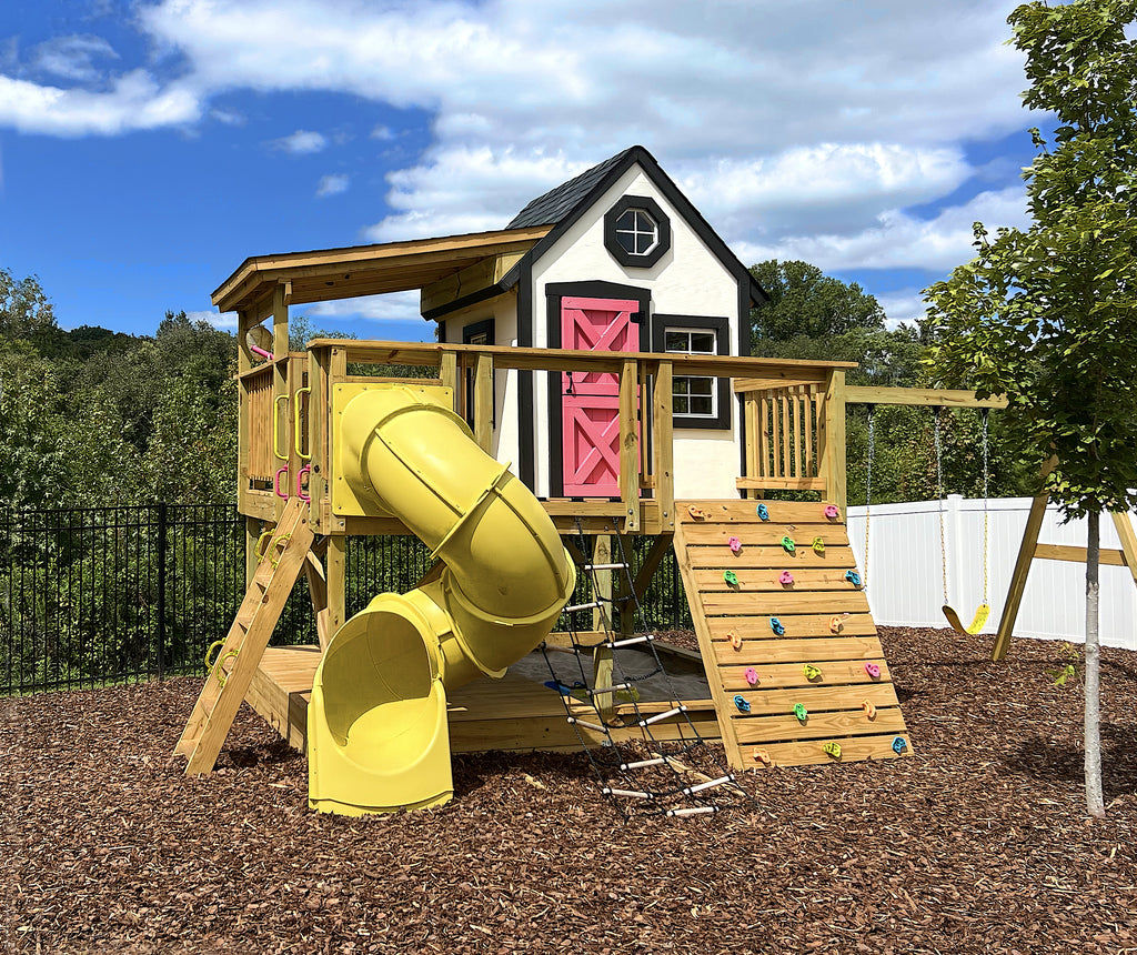 Wooden, elevated playset with pink door, yellow slide, sandbox and climbing wall in a backyard setting.
