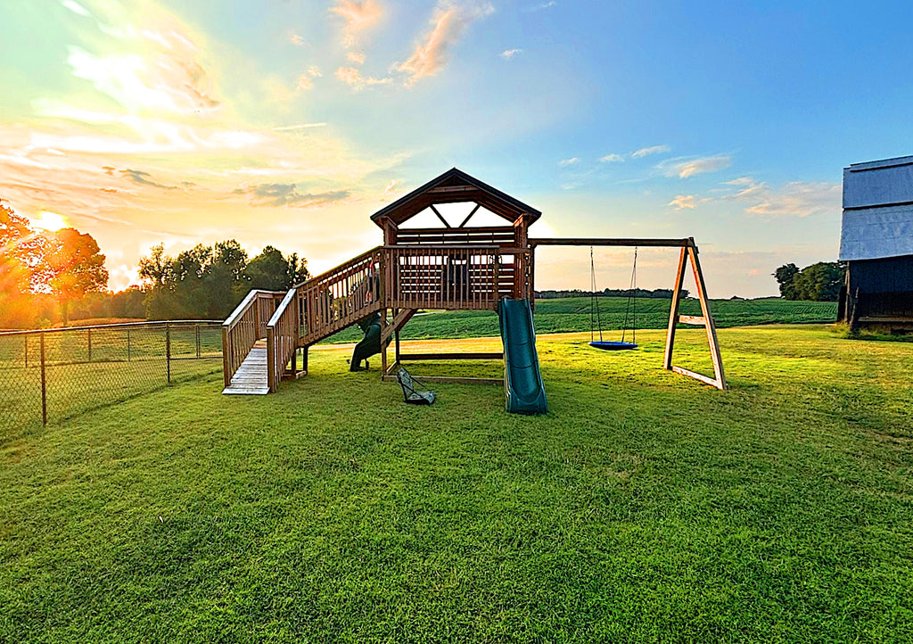 Open gable playhouse on a farm with a setting sun.  The clubhouse has a swingset, slide and ramp.