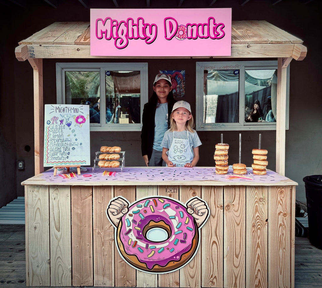 Donut stand with 'Mighty Donuts' sign and two children behind it.