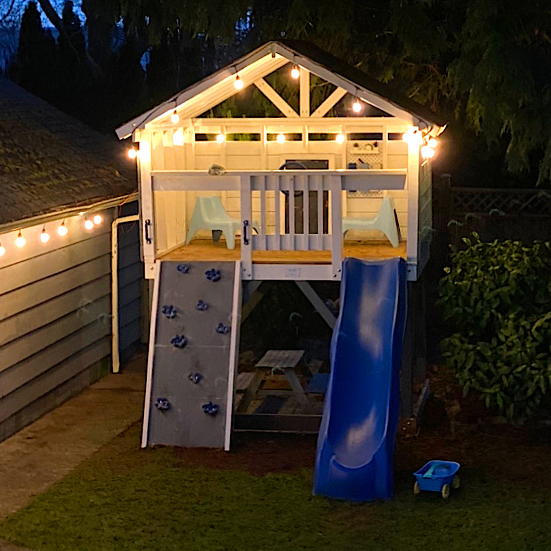Open clubhouse on stils at night with string lighting, a slide and rock wall
