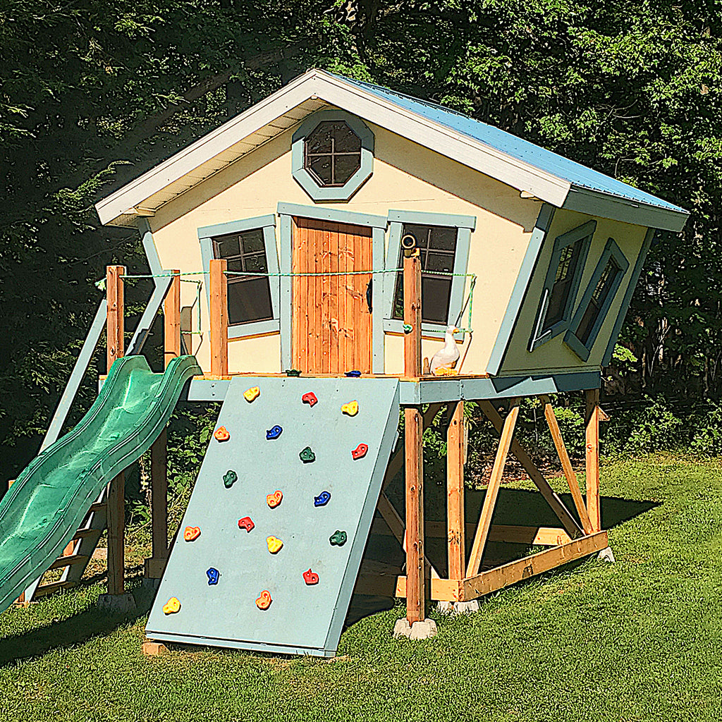 Yellow and blue slanted wall playhouse on stilts with rockwall and slide