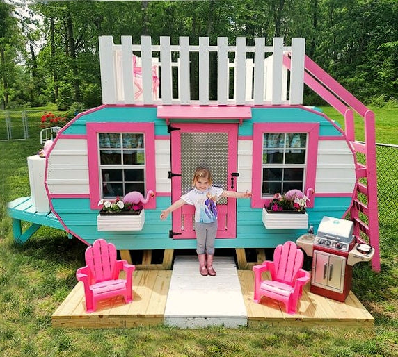 Little girl in front of pink and blue camper playset with chairs, flowerboxes and grill
