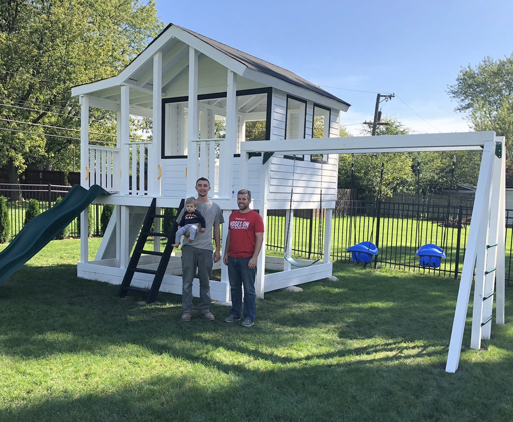 Family standing in front of white, elevated playhouse with swing set